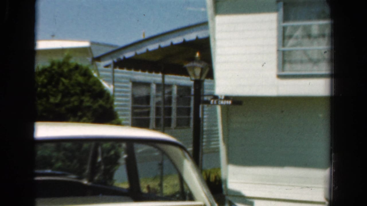 A 1954 California home-movie view of a mobile home exterior with a parked car partly blocking the lower frame.