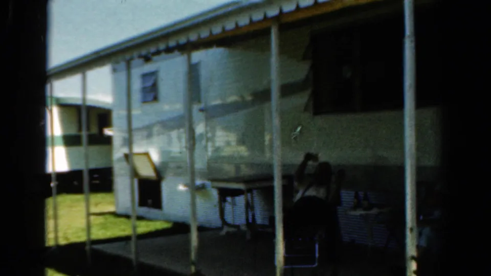 1954 California 8mm frame showing the side of a mobile home, porch posts, lawn, and people seated in the shaded porch area.