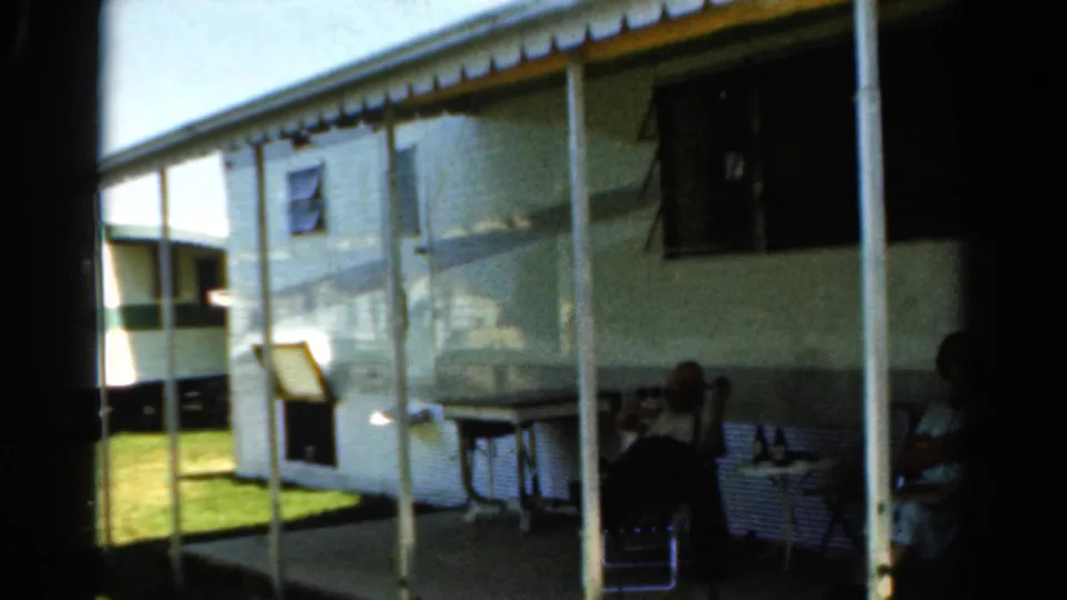 A 1954 California home-movie frame of a mobile-home porch, with people seated in shade beside lawn and porch posts.
