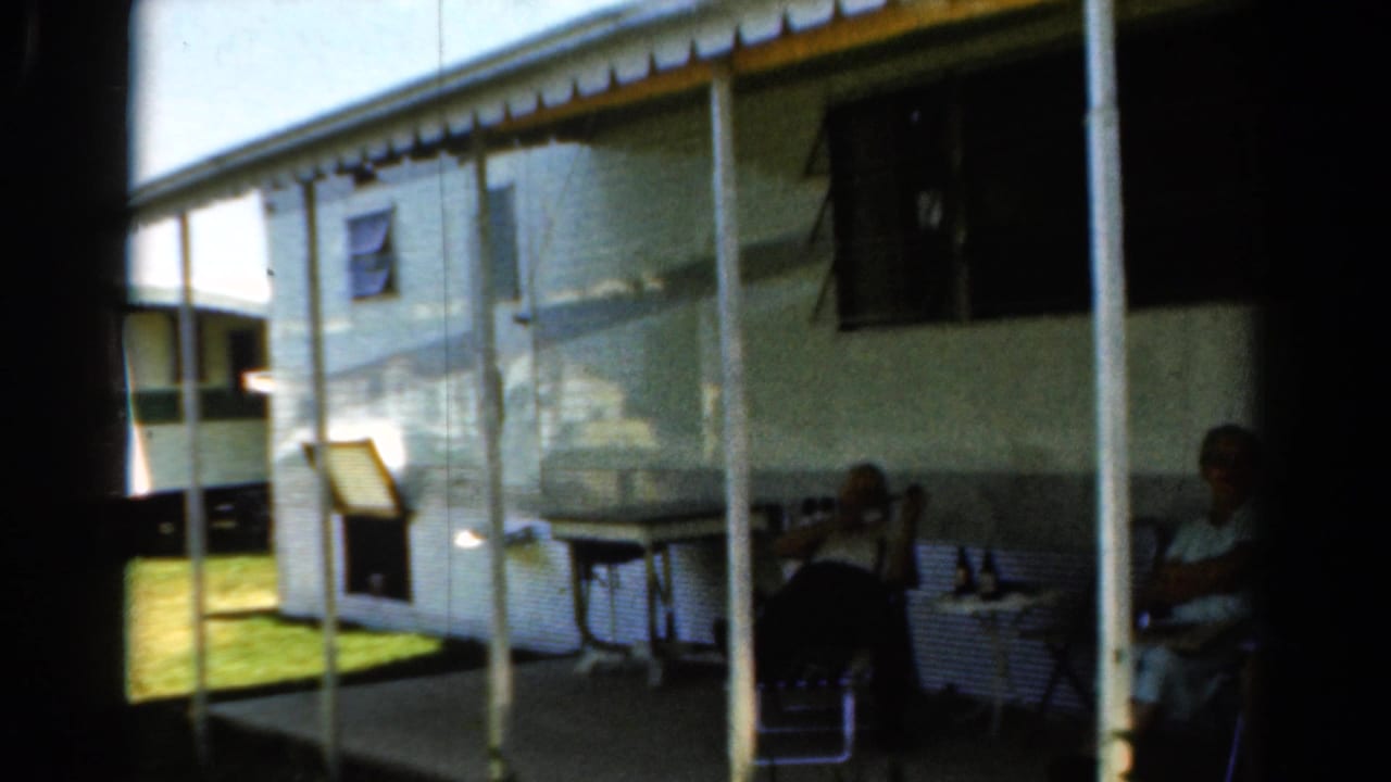 Two adults seated outside a white camper trailer, seen through a dark doorway or screen edge.