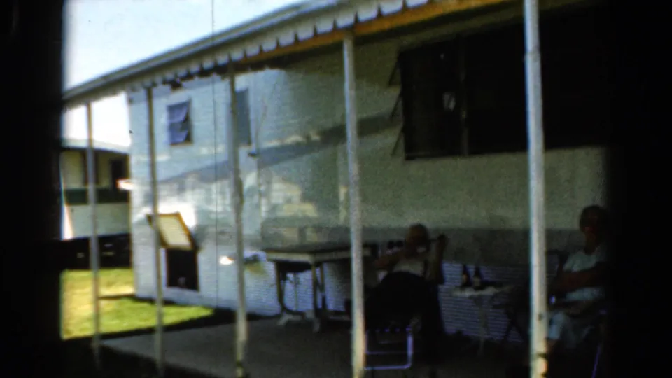 1954 California 8mm frame showing two adults relaxing in chairs beside a white camper trailer with awning poles and a small table.