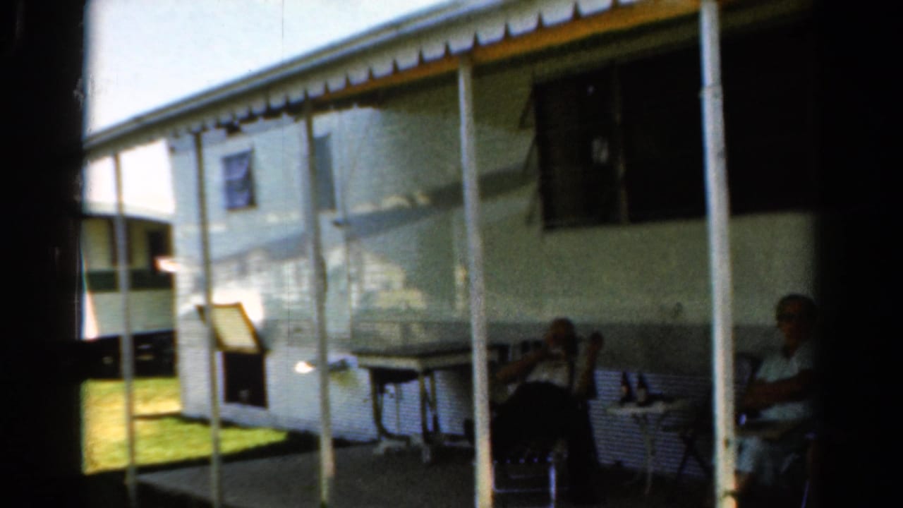 1954 California home-movie frame of adults seated outside a white camper trailer, partly shadowed by the doorway and porch cover.