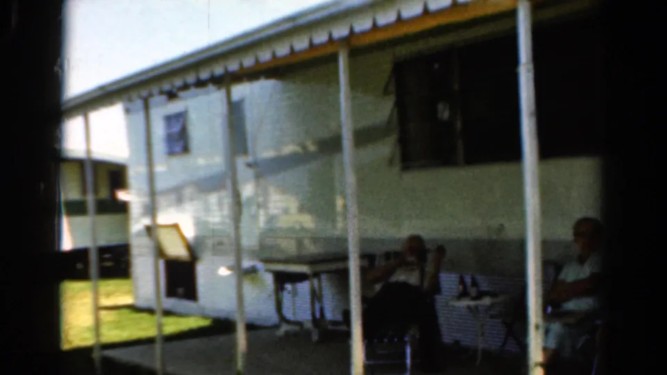 1954 California home-movie frame of adults seated outside a white camper trailer, partly shadowed by the doorway and porch cover.