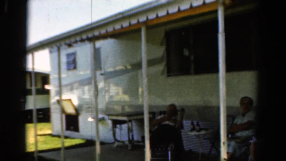 1954 California 8mm frame showing a white trailer exterior, porch poles, lawn chairs, and adults seated in shade.