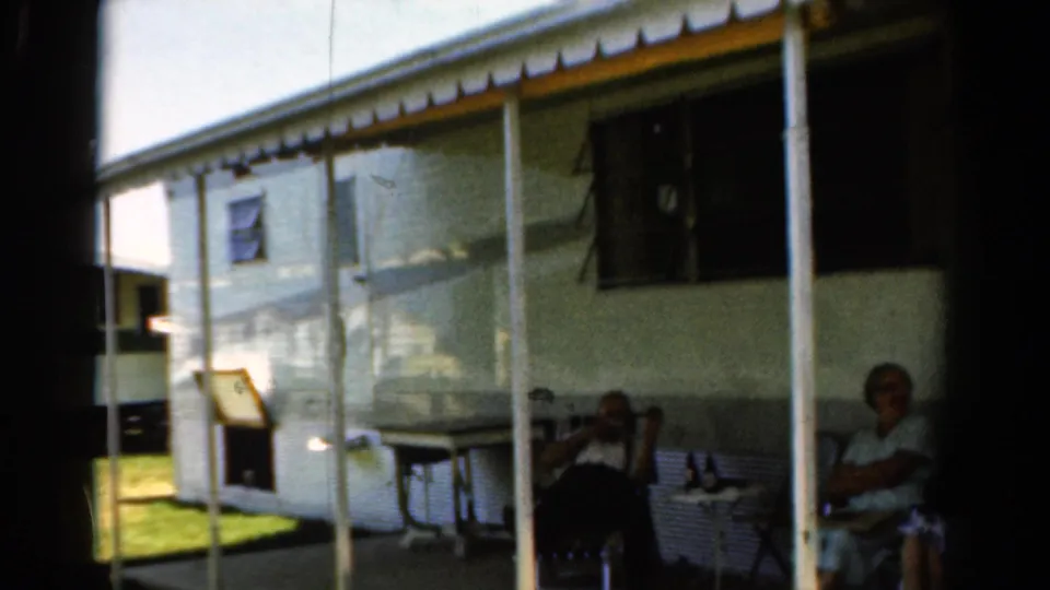 1954 California home-movie frame of adults relaxing outside a white camper trailer, with awning poles, chair seating, and grass visible.