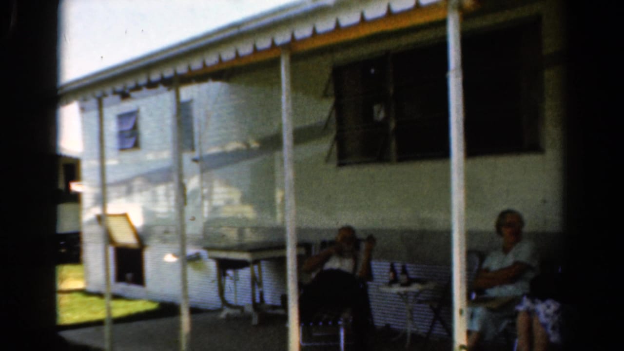 1954 California 8mm frame of older family members sitting on a covered porch with lawn furniture and bottles nearby.
