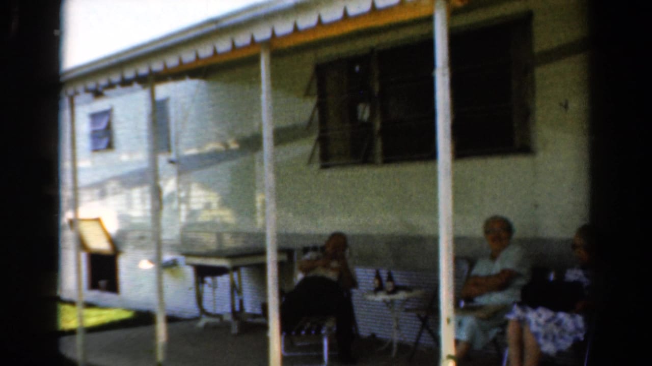 1954 California home-movie frame showing older adults relaxing on a shaded residential porch with mid-century lawn furniture.