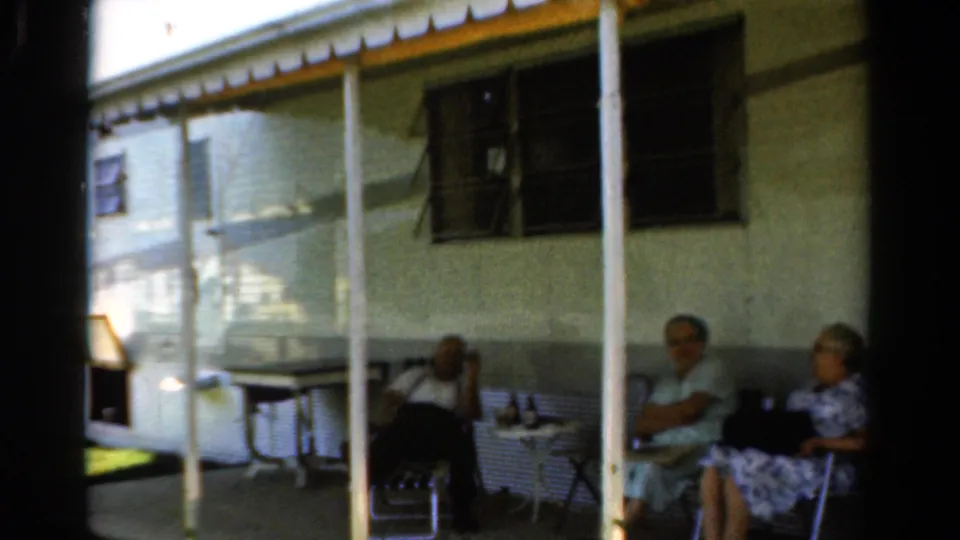 1954 California 8mm frame of family members seated in porch shade near a small table and exterior windows.