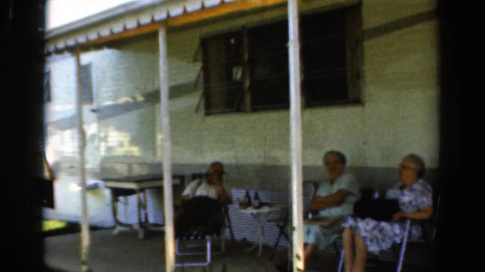 1954 California home-movie frame showing older relatives seated under a porch awning with mid-century chairs and drinks.