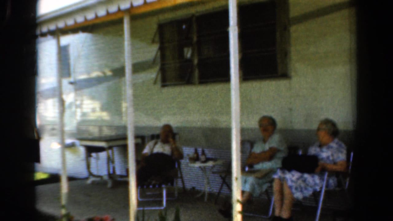 Three older women sit on a shaded porch beside a small table and folding chairs.