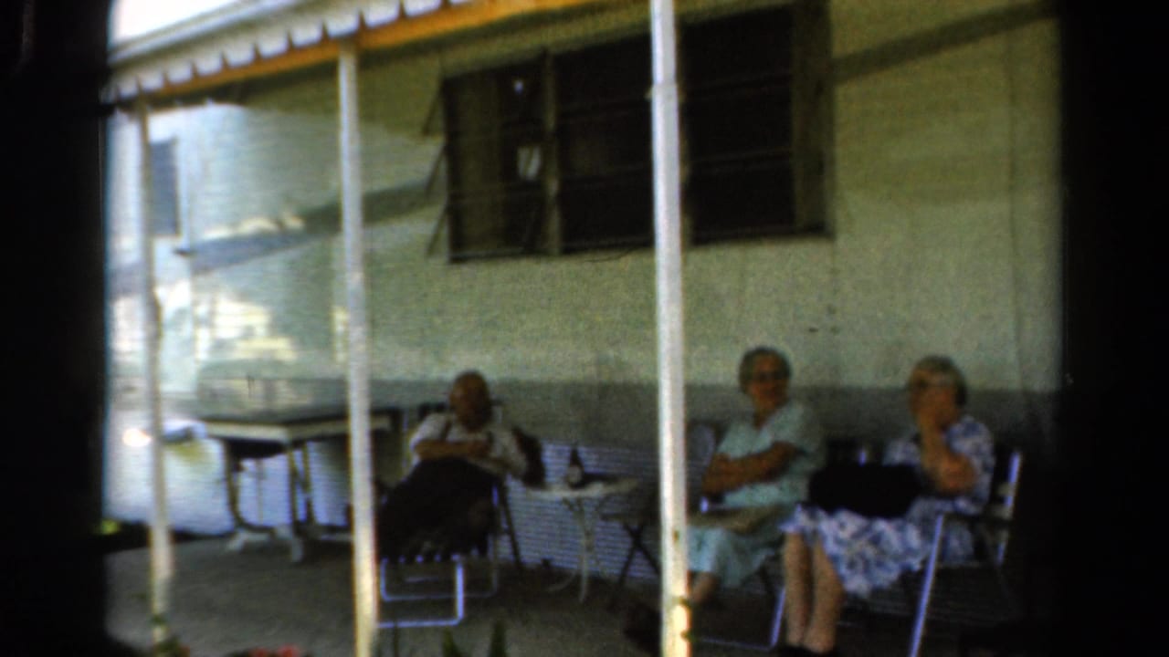 In 1954 California home-movie footage, three older women sit on a shaded porch with folding chairs and a small table visible.