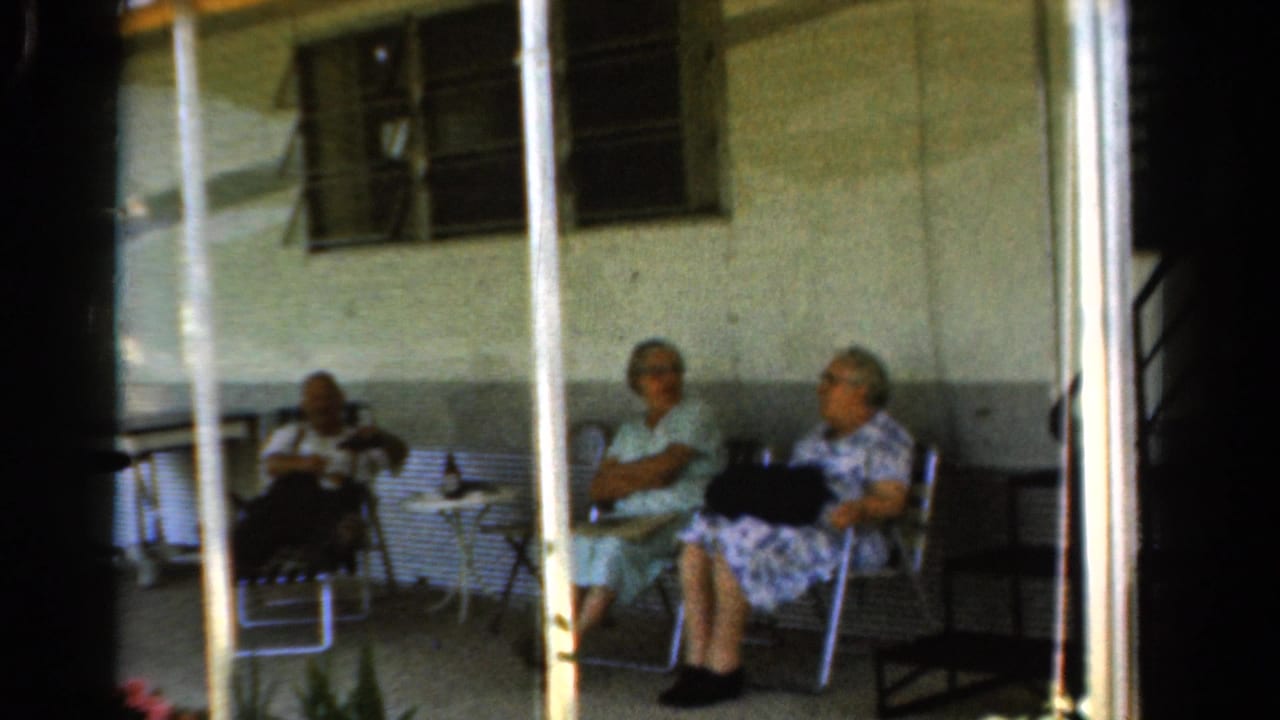 Three older adults sit on a covered porch with chairs, a small table, posts, and windows behind them.