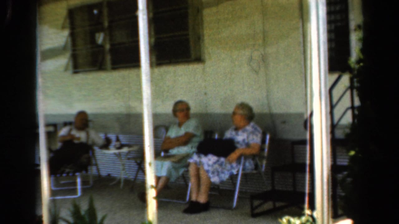 Two elderly women sit in metal chairs on a porch while a third person is partly visible at left.