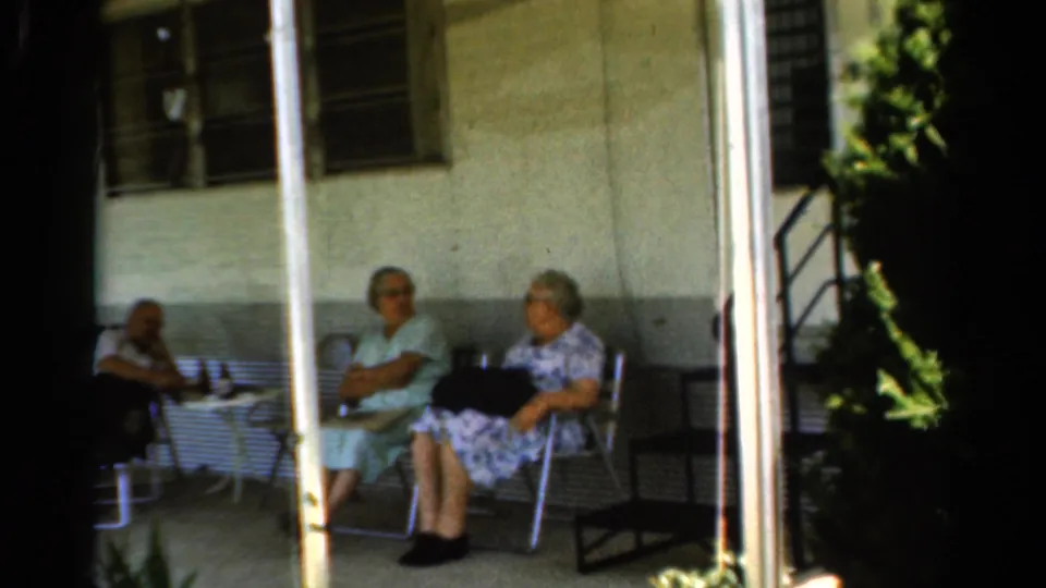 1954 home-movie frame of two elderly women seated in lawn chairs on a covered porch, with the third seated adult only partly in view.