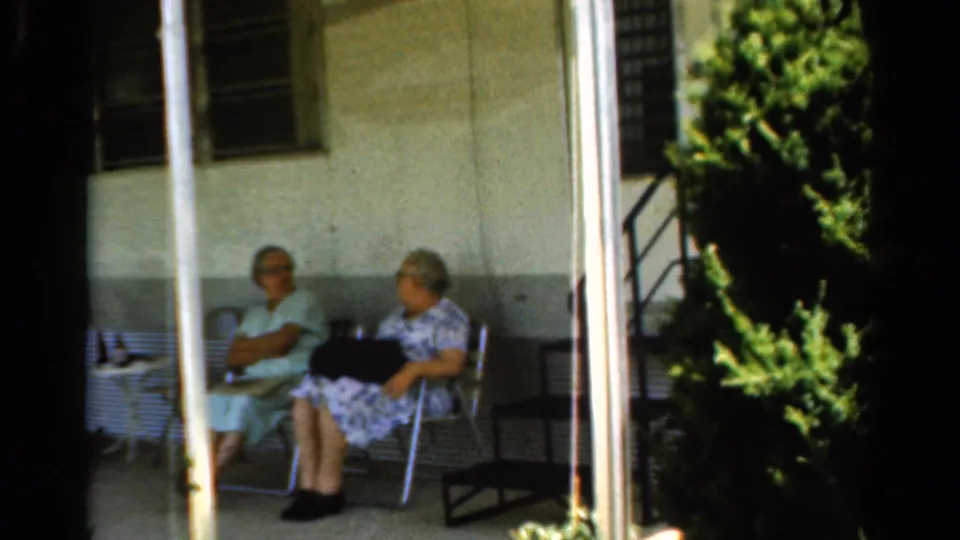 Two elderly women remain seated on a shaded porch in 1954 California footage, framed by a white post, wall, window, and garden foliage.