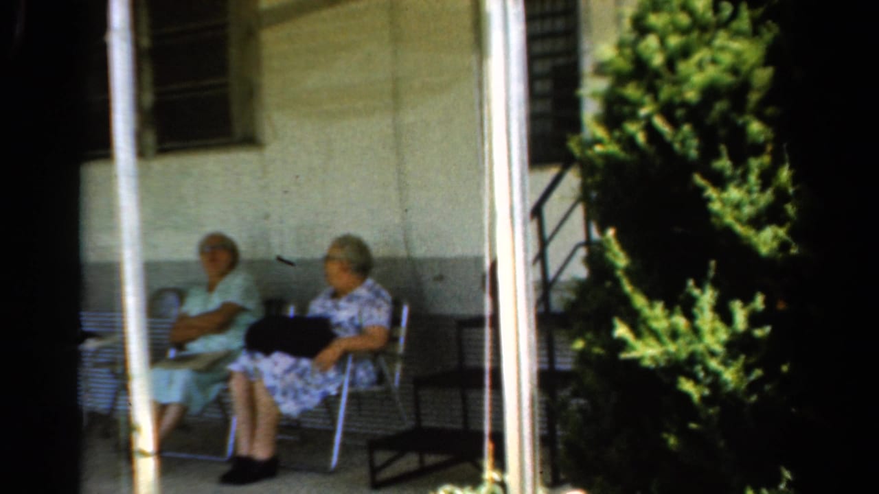 Two elderly women sit in folding chairs on a shaded porch beside a large shrub.