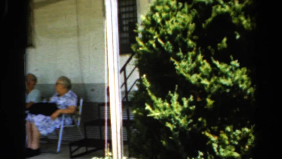 Two elderly women in light dresses sit on a porch next to a dense evergreen shrub in 1954 California home-movie footage.