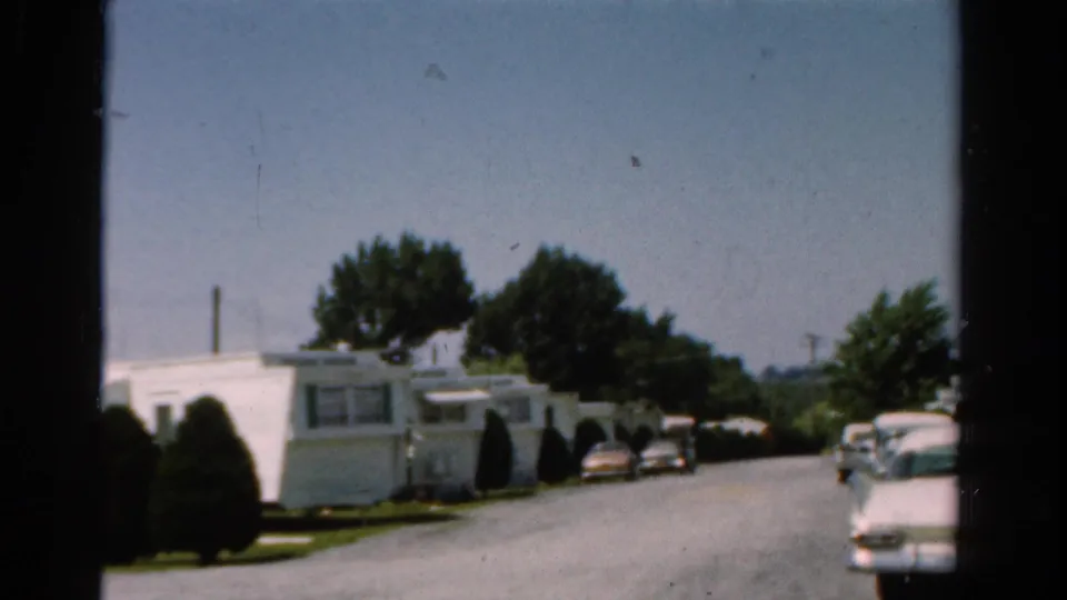 1954 California 8mm frame showing white mobile homes, green shutters, trees, and parked cars on a quiet residential road.