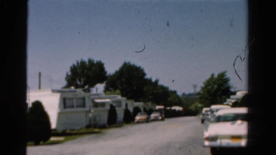 1954 California street view with trailer homes, clipped shrubs, parked cars, and a white sedan partly entering the foreground.
