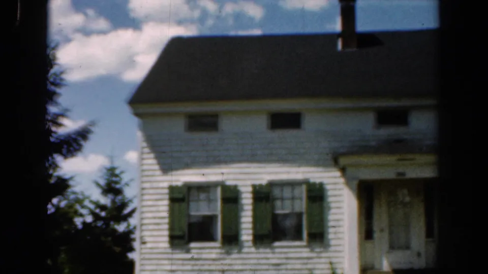 1954 California home-movie frame of a white two-story house with green shutters, a dark roof, windows, and front entry.