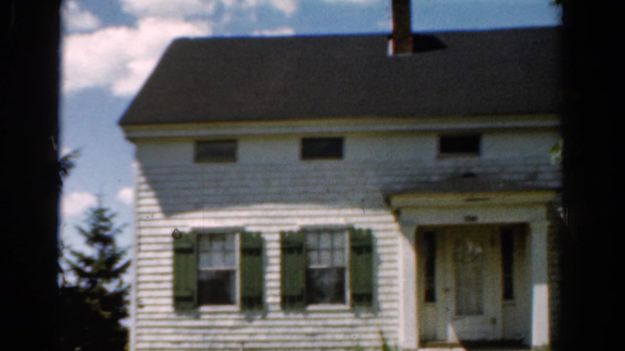 1954 California home-movie frame showing a white two-story farmhouse with green shutters, a dark roof, chimney, and trees at the edges.