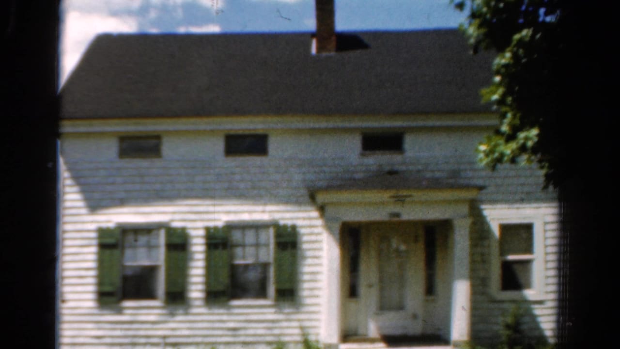 1954 California exterior view of the farmhouse facade, showing shuttered windows, a central doorway, roofline, chimney, and surrounding foliage.