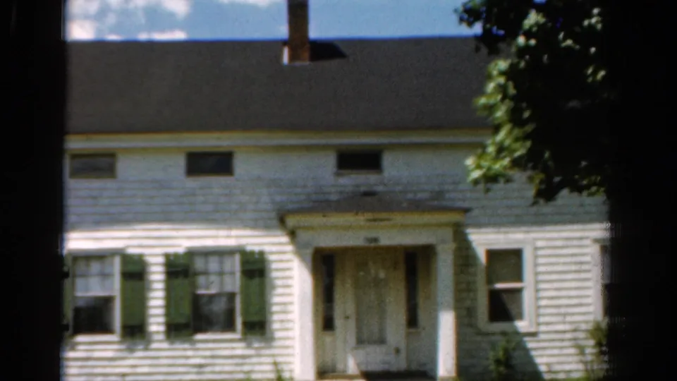 1954 California 8mm frame with a slightly lower view of the white farmhouse, showing shuttered windows, front door, roof, and leafy branches.