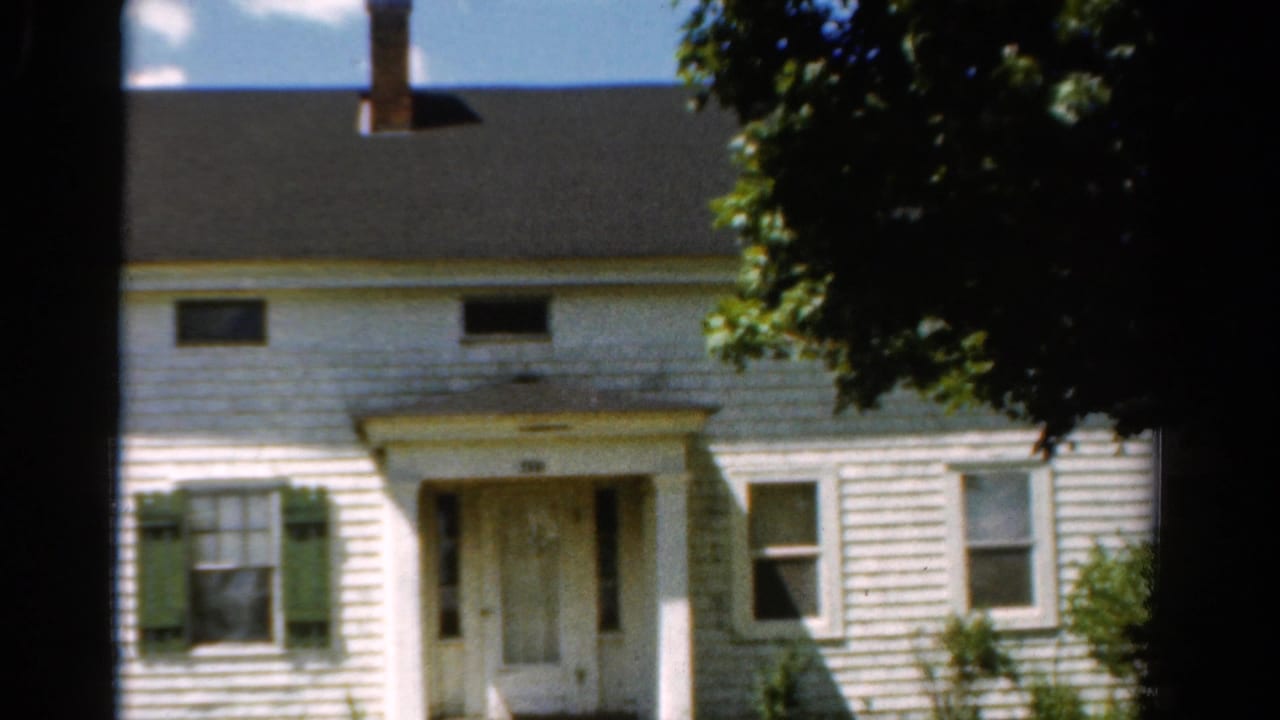 1954 California 8mm frame showing a white two-story clapboard house with green shutters, porch columns, shrubs, and tree shade.