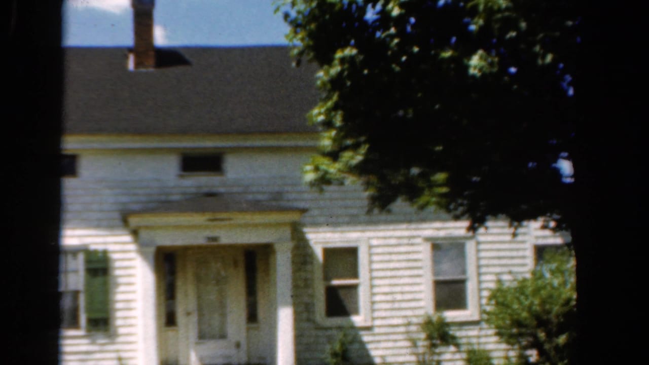 1954 California home-movie frame of a white clapboard residence with green shutters, a central doorway, porch roof, and tree canopy.