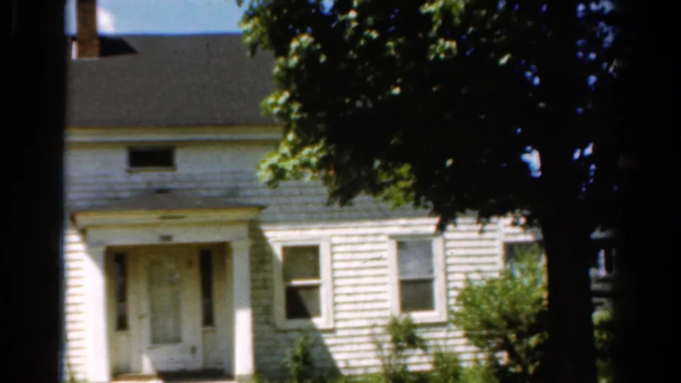 1954 California 8mm frame showing the front entrance of a white house, green shutters, shrubs, and a dark tree trunk crossing the view.