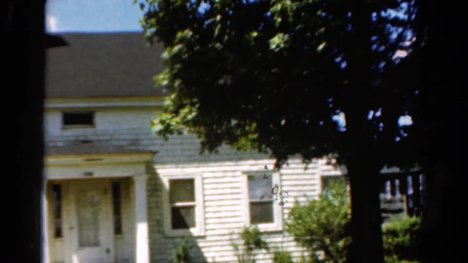 Final 1954 California frame of the pan, showing the white house exterior, green shutters, front door area, shrubs, and heavy tree shade.