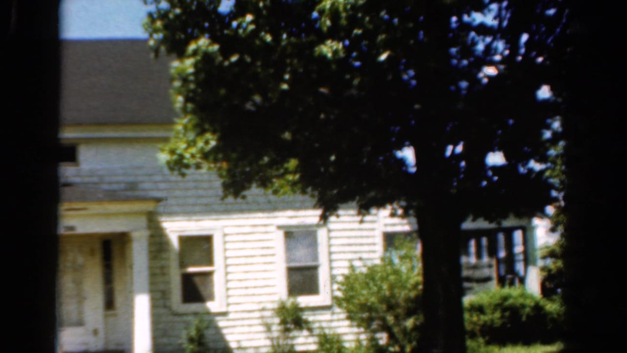 1954 California home-movie frame with a white house behind a dark tree trunk, leafy canopy, porch opening, windows, and low shrubs.