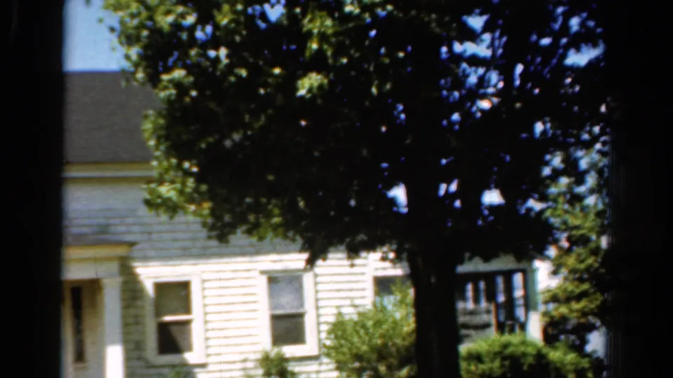 1954 California 8mm frame showing the side of a white clapboard house, two windows, shrubs, blue sky, and dense tree shade.