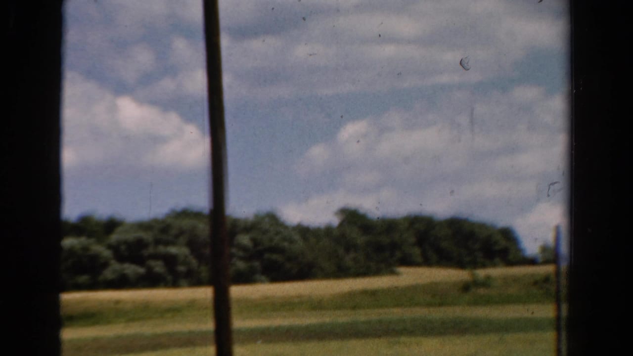 1954 California 8mm frame showing rolling green fields, a distant treeline, and blue sky viewed through dark window framing.