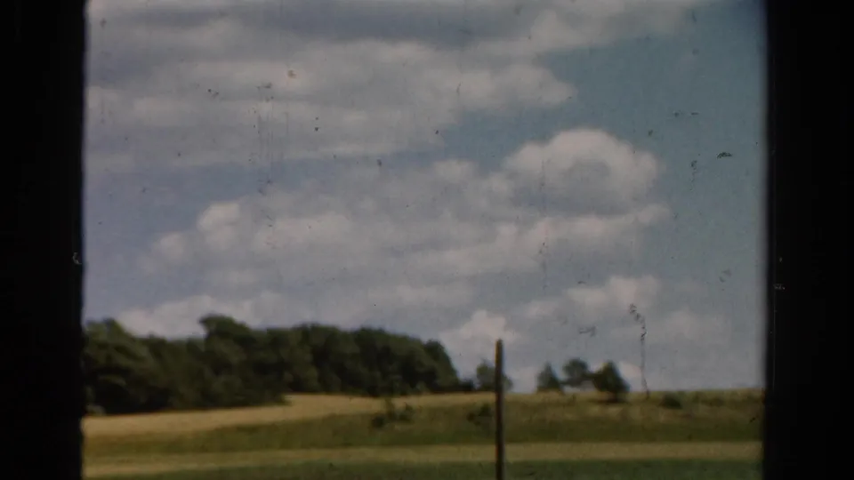 1954 California 8mm frame with green fields, distant trees, a narrow pole, and bright clouds viewed from behind a dark window edge.