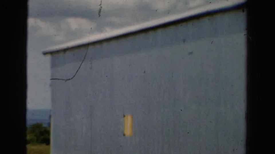 1954 California 8mm frame showing a plain light-blue building wall, dark window framing, and a glimpse of sky and landscape beyond.