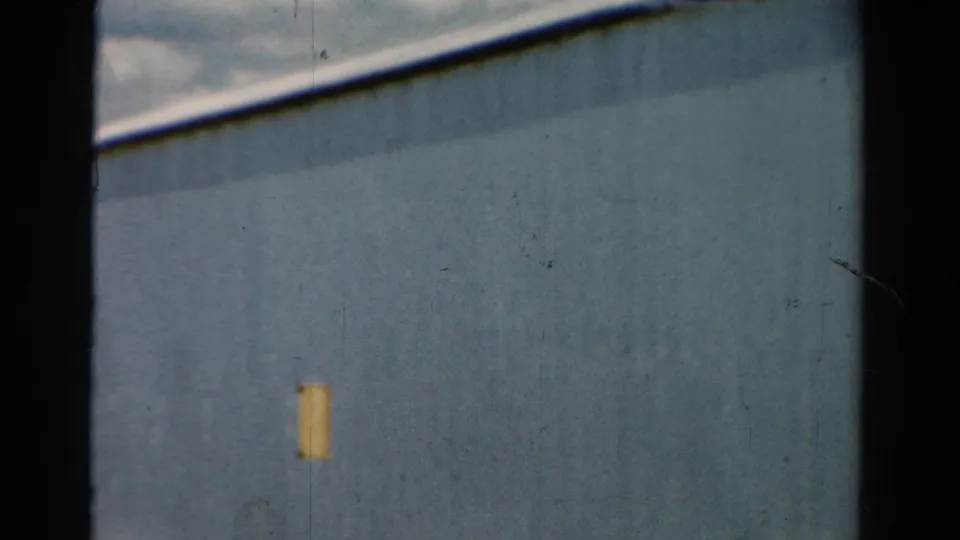 1954 California 8mm frame with blue metal siding, a small yellow wall patch, muted clouds, and a dark vertical border through the shot.