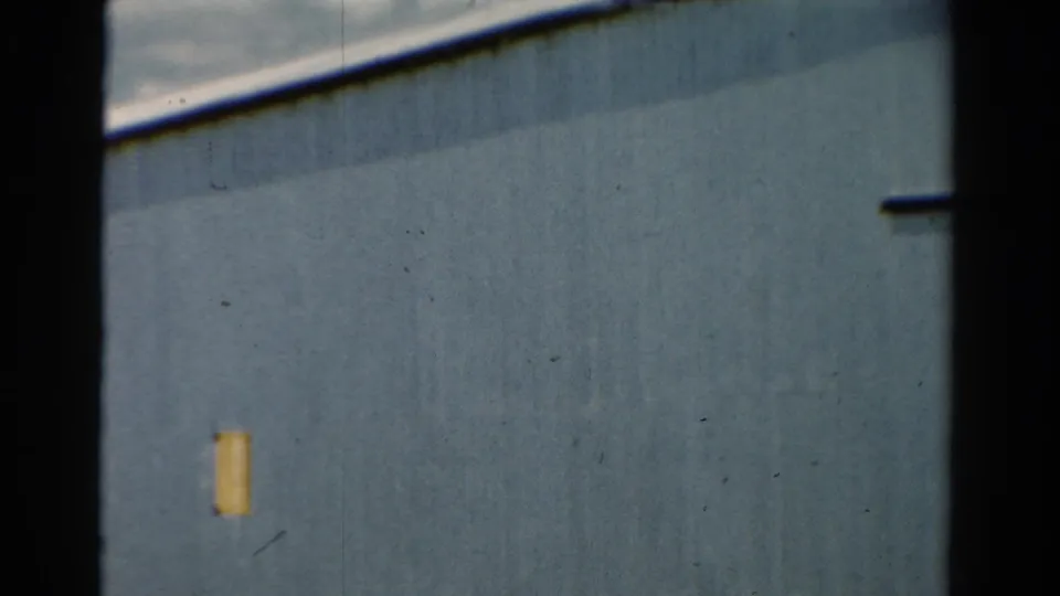 1954 California 8mm frame showing a close, slightly soft view of blue metal siding, yellow patch, upper roofline, and dark film border.