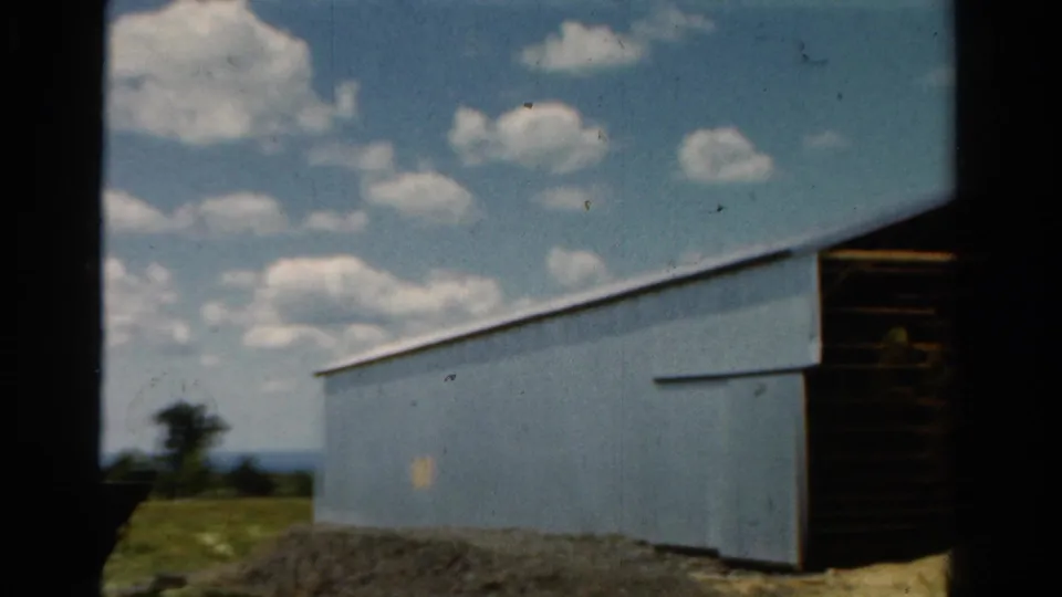 1954 California 8mm frame showing a wider rural view of the blue outbuilding, scattered clouds, grassy ground, distant tree, and dark side opening.