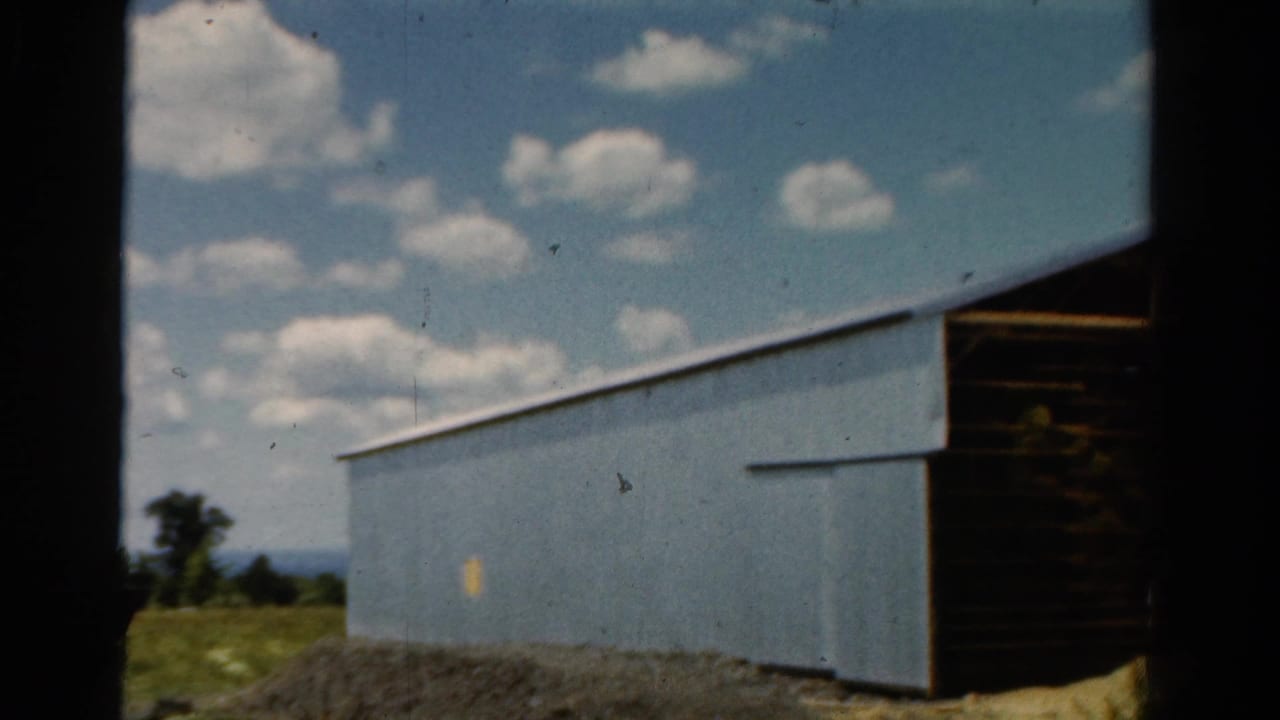 1954 California 8mm frame showing a pale blue barn under scattered clouds, with a dark open opening at the right edge.