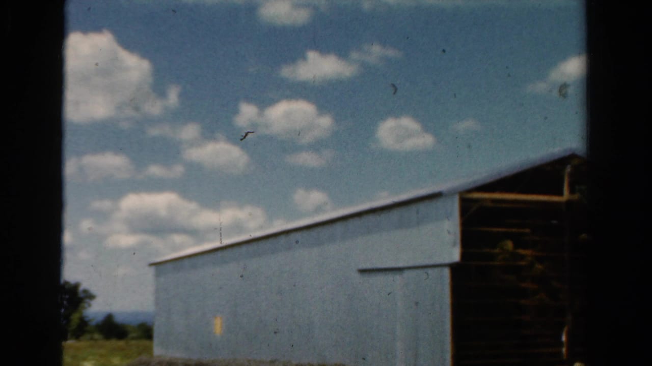 1954 California home-movie frame of a blue barn exterior, dark open doorway, grassy edge, and bright clouded sky.