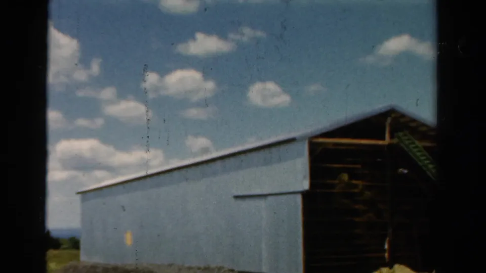 1954 California 8mm still with the blue barn wall closer in view, a shadowed opening, and scattered clouds above.