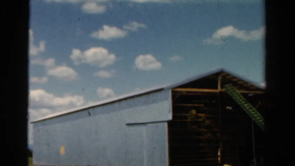 1954 California frame showing the blue barn, a dark open bay, and a green ladder leaning against the structure below scattered clouds.