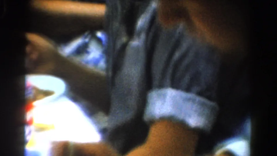 1954 California home-movie frame showing an adult at a table with rolled sleeves, food items, and a bright tabletop area.