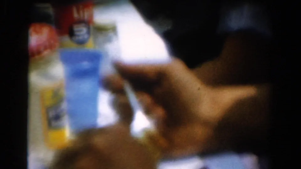 Soft 8mm frame from a 1954 California indoor meal, with a hand holding a cup near a blue container and packaged food.