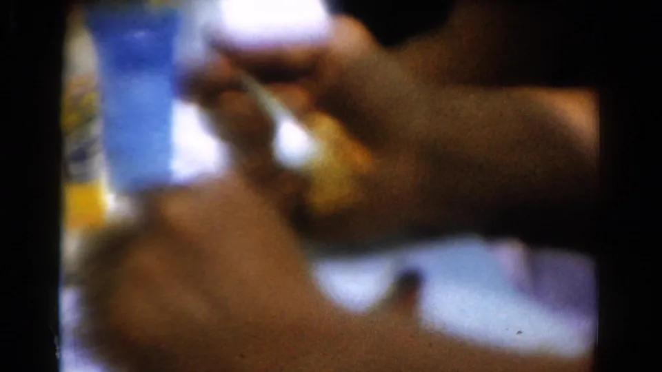 1954 California 8mm frame of a hand holding a cup at a dim indoor table, with packaged items blurred in the background.