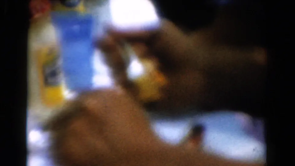 1954 California 8mm frame showing a hand with a fork near a plate, with a blue bottle and yellow container in soft background focus.