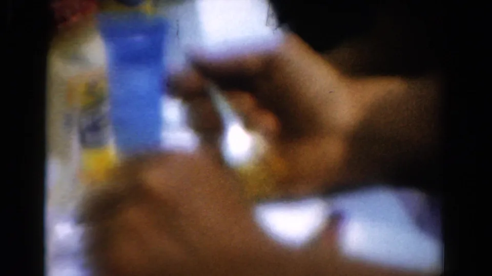 Close 1954 California home-movie frame of a hand and fork beside a plate, with colored tabletop items blurred behind.