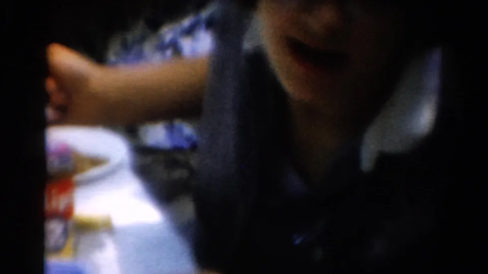 1954 California home-movie frame showing part of a person at a table, with an arm extended near a plate and tabletop items.