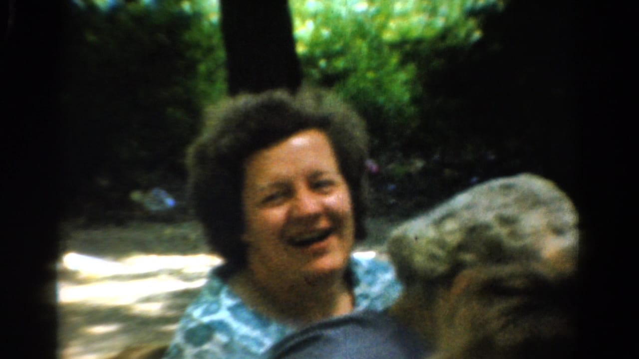 A smiling woman with curly dark hair faces the camera in 1954 California home-movie footage, with trees and a large stone form nearby.